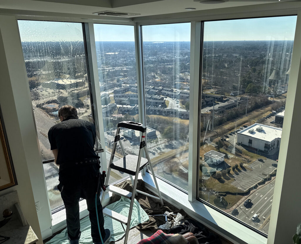 Taken with UploadCam • Skyline Tinting LLC Worker on a step ladder inside a high-rise, cleaning large glass windows with water droplets, city streets and ocean view outside