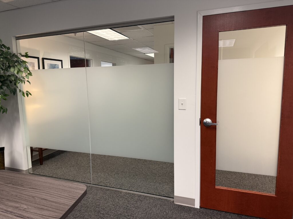 Glass-walled office partition with frosted lower panels, a wooden door on the right, and a potted plant to the left.