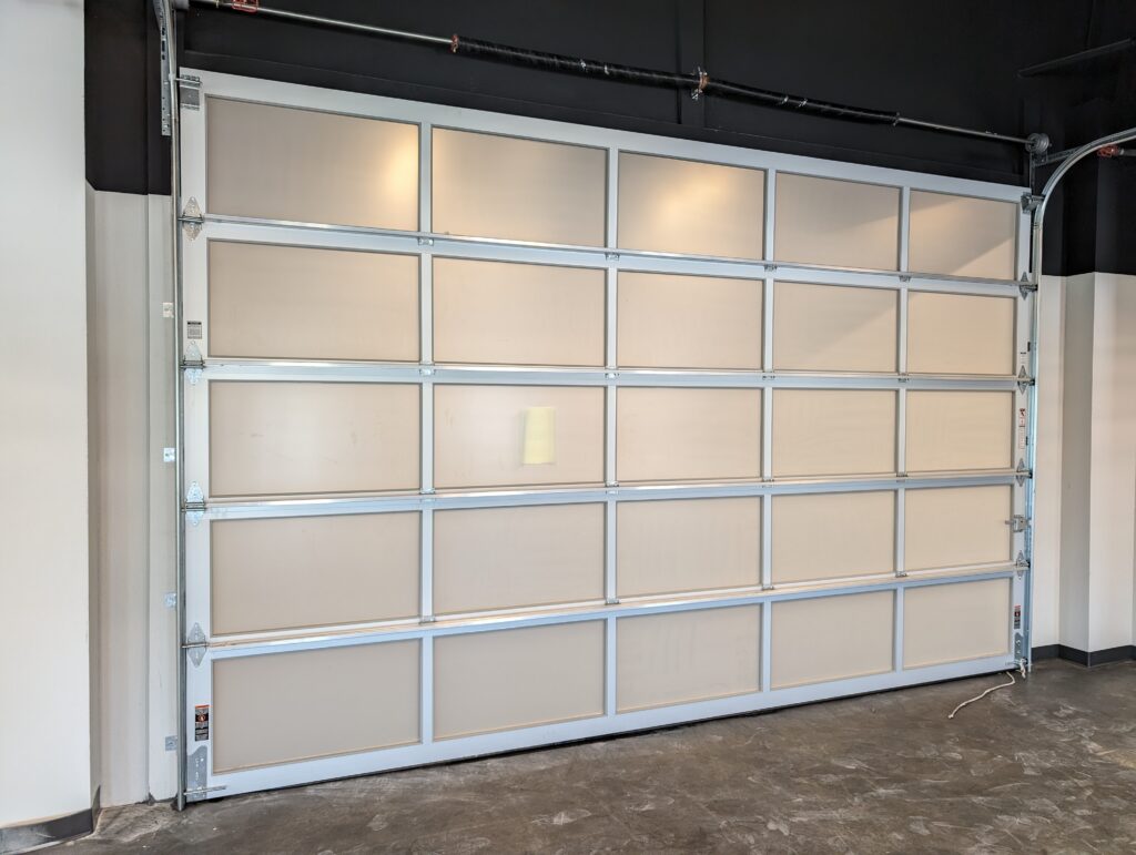 PXL_20240528_191140378 • Skyline Tinting LLC Industrial sectional garage door with metal frame and beige panels in a warehouse interior. overhead cables visible.