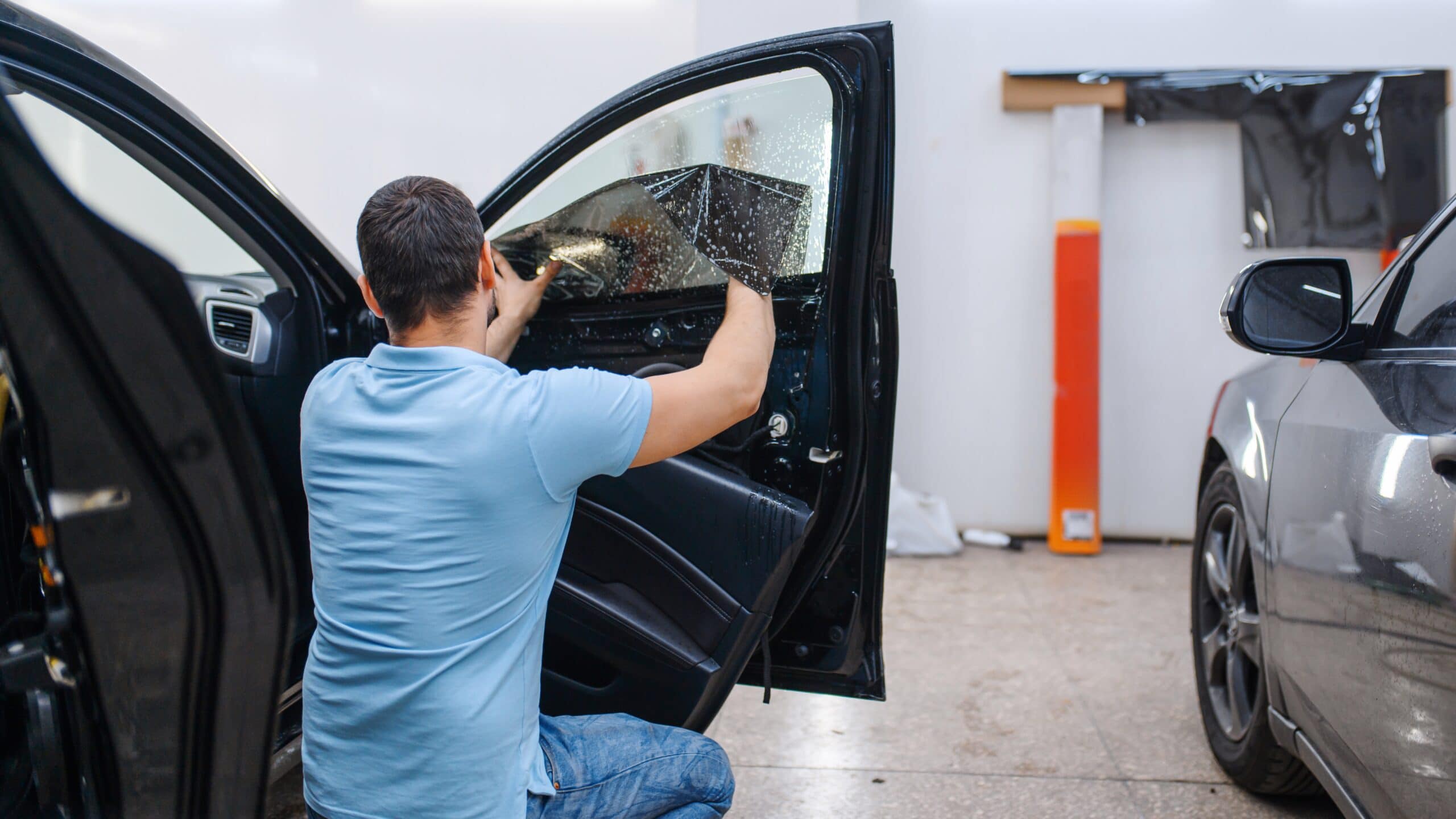 Man in a light blue polo kneels beside an open car door, applying window tint film to the side window inside a workshop.