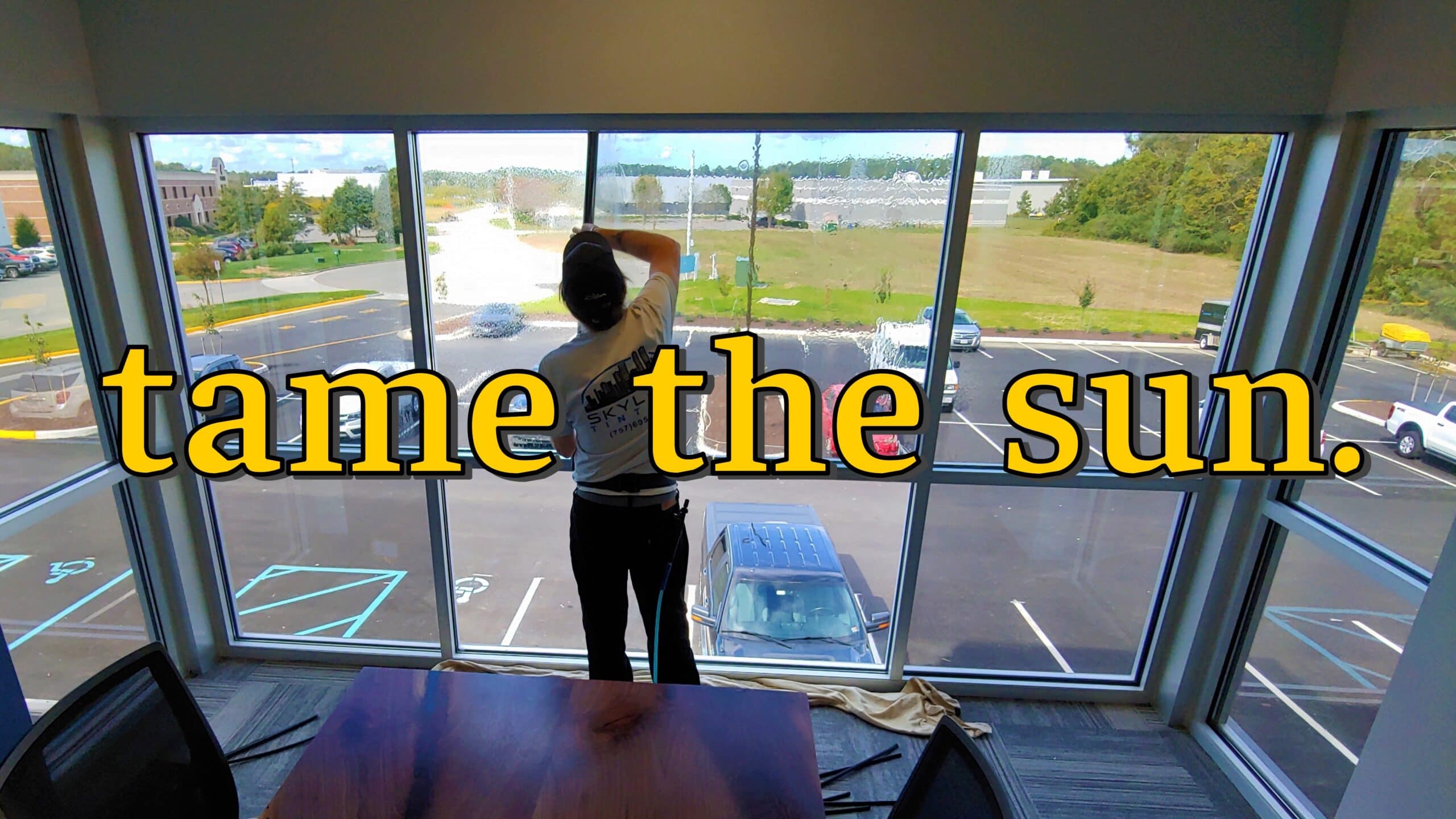 Indoor scene of a person cleaning a large glass window with a squeegee; parking lot and trees visible outside, with bold yellow 'tame the sun' text across the image.