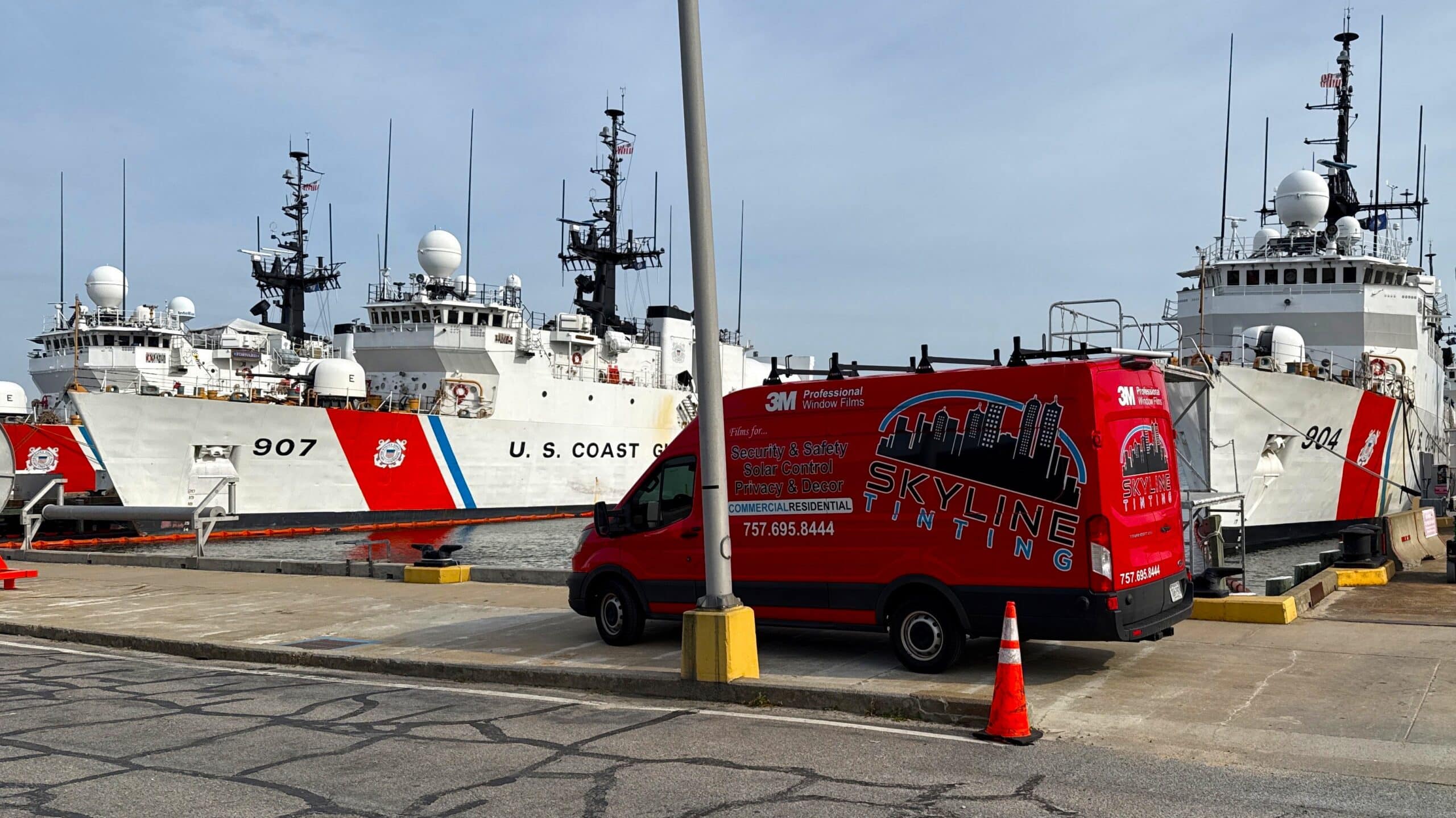 Two U.S. Coast Guard ships docked at a pier, with a red Skyline Window Tinting van parked in front.