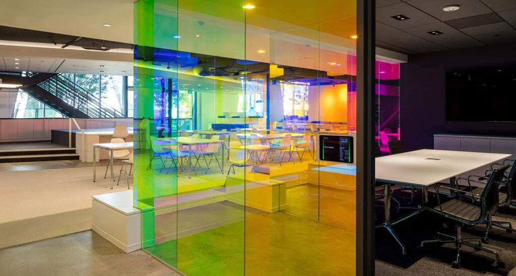 Open workspace with rainbow-colored glass panels forming a partition around a meeting area with tables and chairs visible behind the glass.
