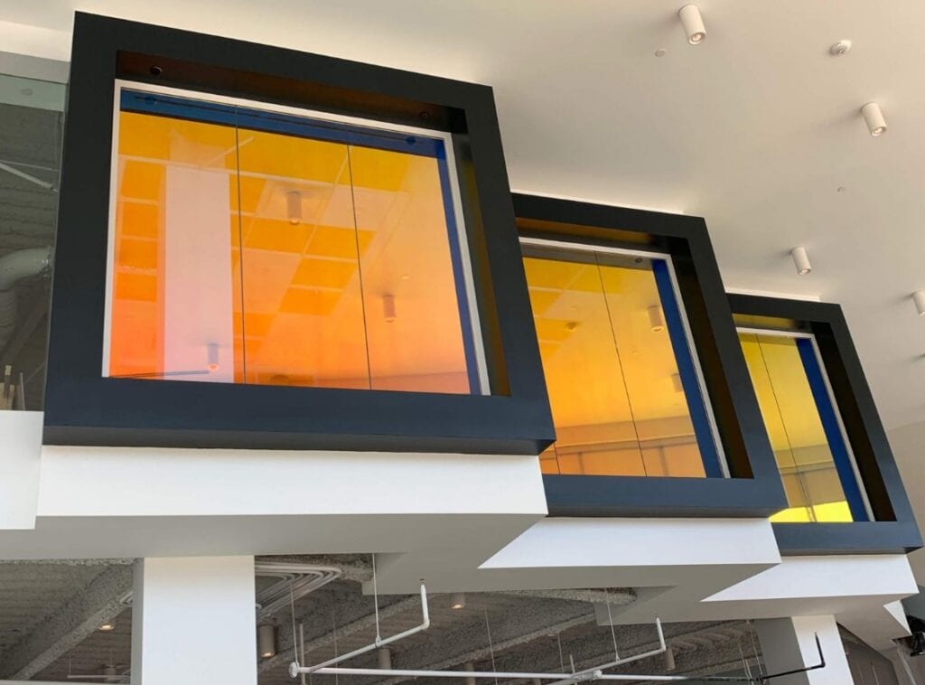 Three large black-framed square windows with orange–yellow tinted glass in a modern interior atrium or lobby edges above white beams.